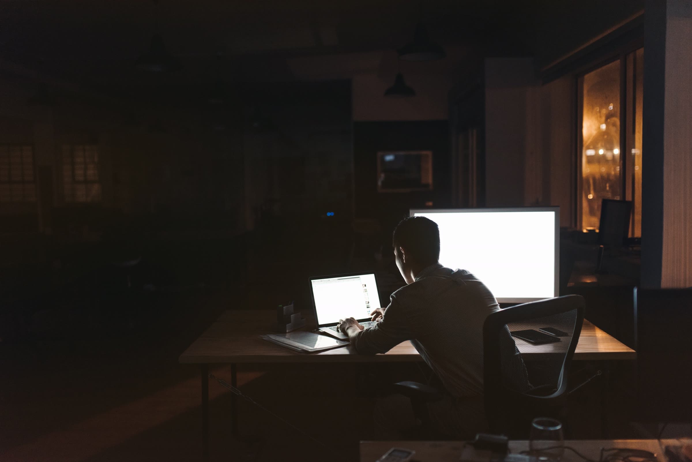 A man working alone at a desk at night, lit by a laptop and monitor in a darkened office.